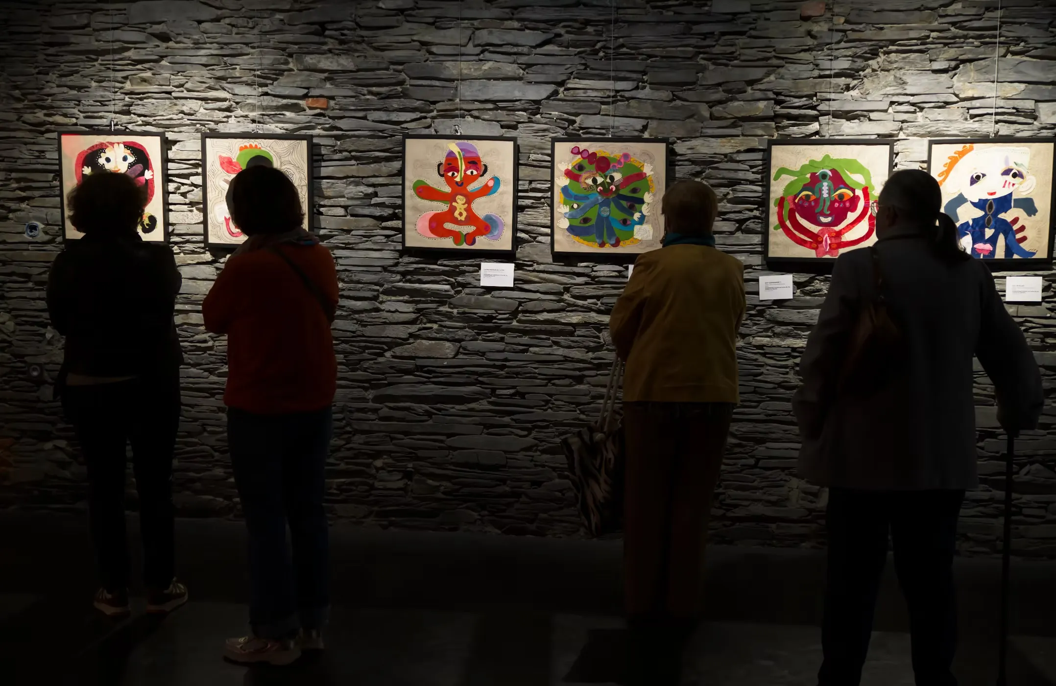 Visiteurs en silhouette devant des portraits en tissus assemblés sur mur en pierre, exposition Katherine Roumanoff Trélazé